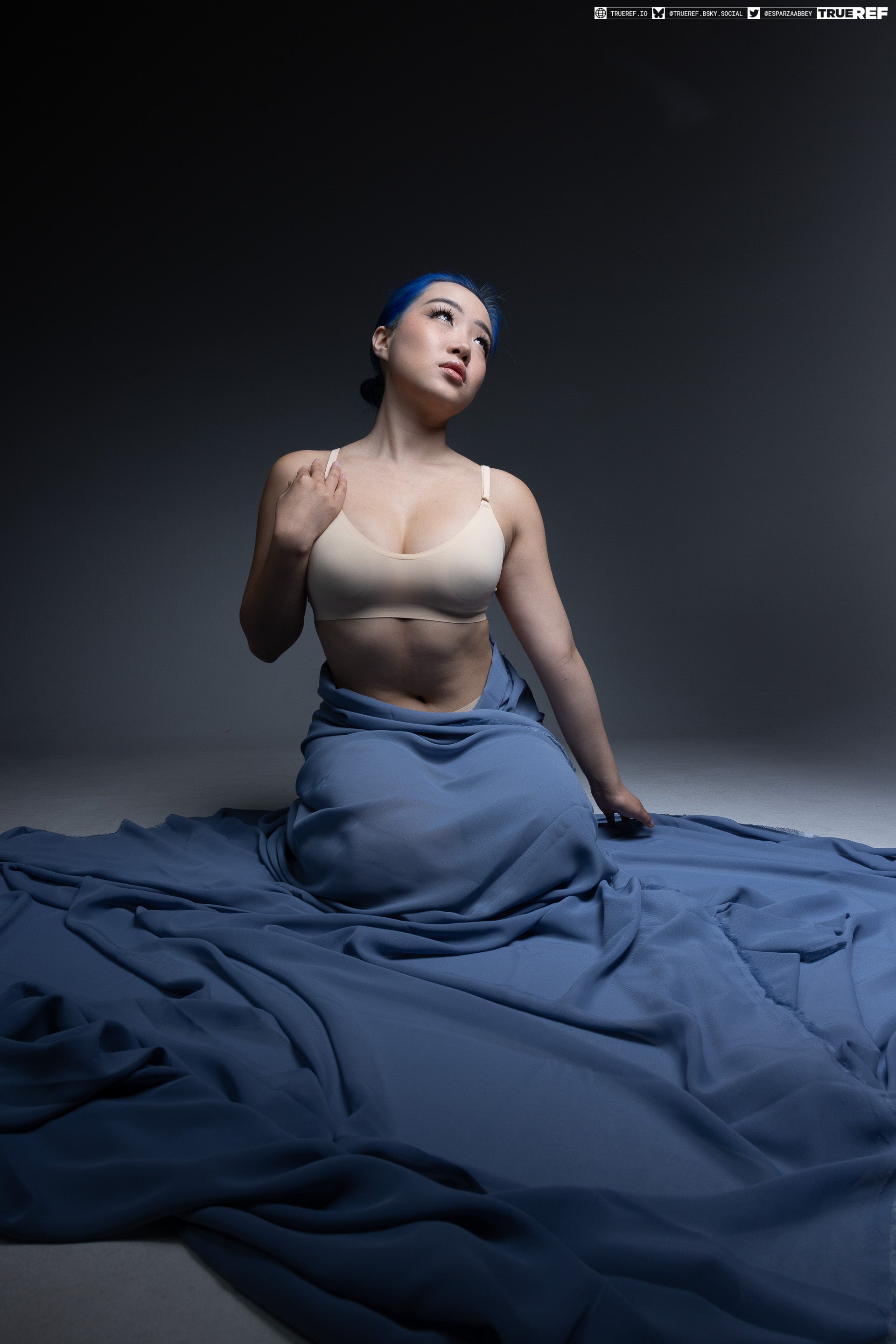 a woman with blue hair is sitting on the floor draped with a large blue sheet, wearing a bra and looking up. the image is a studio shot with a gray background.