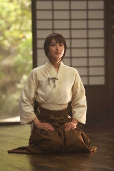 a woman wearing a white kimonostyle top and brown hakama pants kneels on a dark wood floor. she is facing to the left of the frame with her hands resting on her lap. behind her is a light wood and opaque sliding door. the lighting is natural and appears to be coming from the sliding door. the overall mood of the image is peaceful and serene.