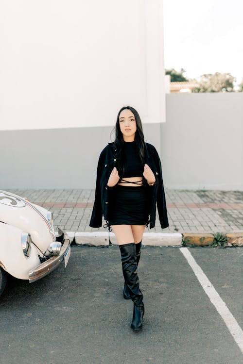 a young asian woman stands confidently in a parking lot, posing for a photo. she is wearing a stylish black outfit consisting of a mini dress, a black denim jacket, and thighhigh black boots.  she's standing in front of a vintage white volkswagen beetle. the background includes a modern white building and some greenery. the overall style is fashionable and chic.