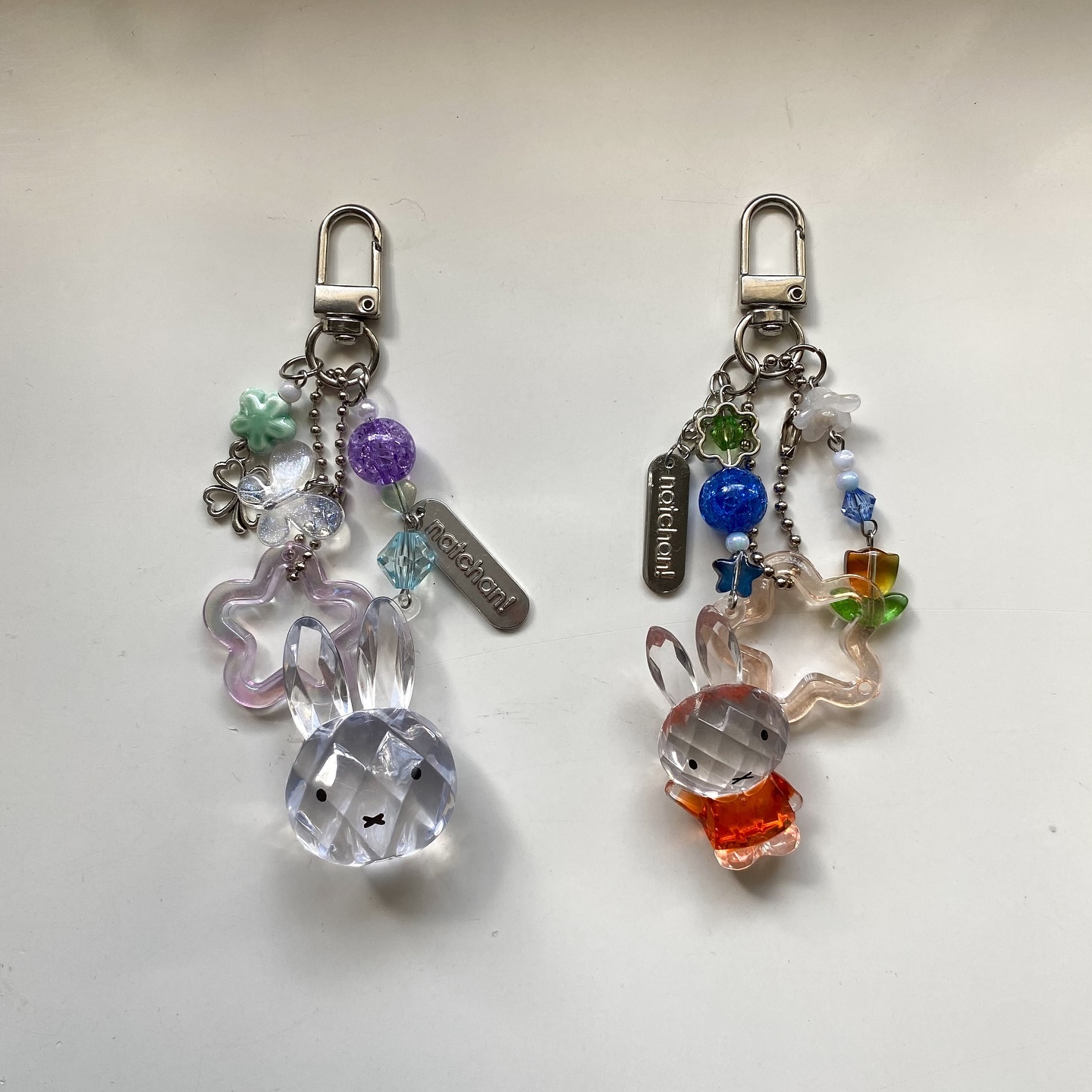 the image features two handmade keychains, each adorned with a clear rabbit charm, a starshaped translucent charm, metal and glass beads, and a keychain clip. the keychain on the left has a clear rabbit, a clear star with pink edges, a purple bead, and a tag. the keychain on the right has a rabbit with an orange body, a clear star with rainbow edges, and a blue bead. both keychains have the same silver clover charm.