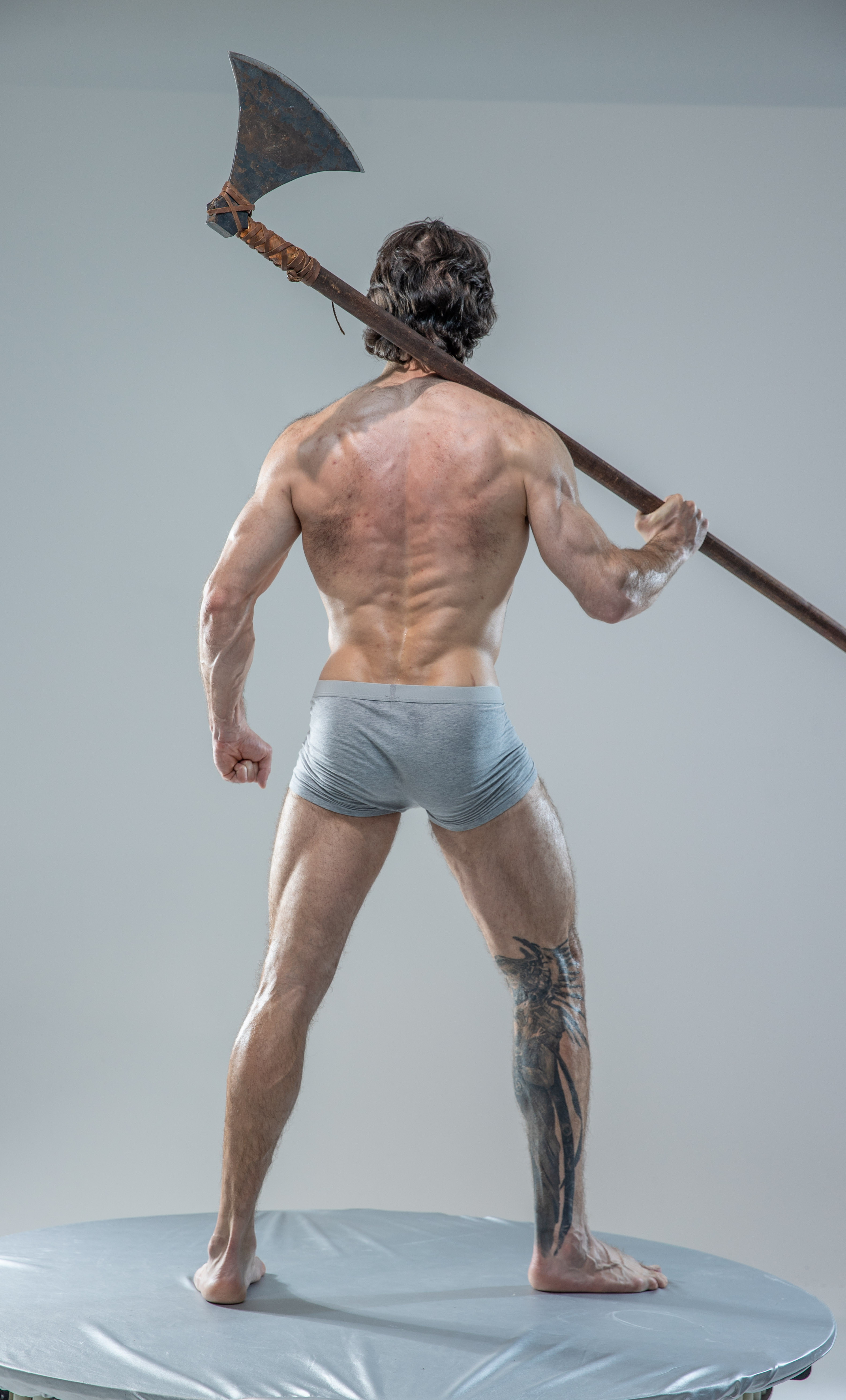 a muscular man stands with his back to the camera, showcasing his physique. he holds a large, worn axe over his shoulder.  he is shirtless, wearing only grey underwear and displaying a tribal tattoo on his left leg. the setting appears to be a photography studio with soft, even lighting.