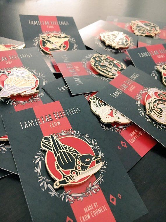 the image shows a collection of "familiar feelings" enamel pins displayed on their backing cards. the pins feature stylized illustrations of animals like a crow, snake, owl, frog, and newt, each holding a weapon or item. the cards have a black background with a red stripe, displaying the pin's title and the text "made by crow council" at the bottom.