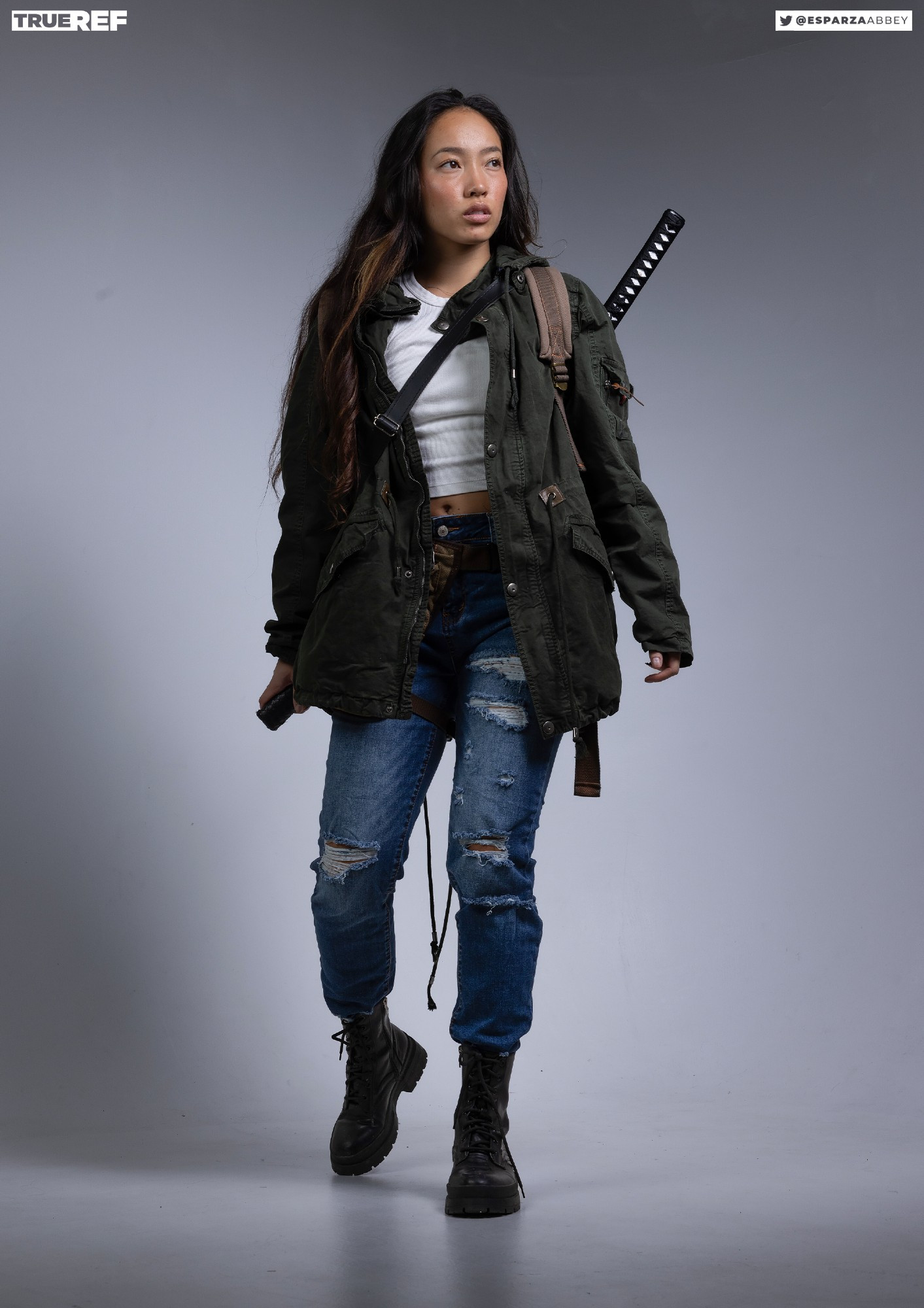 a fullbody shot of a young woman, model esparza abbey, posing against a grey background. she's wearing a green jacket, ripped jeans, black combat boots, a backpack, and carrying a katana on her back. she looks away from the camera, showcasing a casual yet stylish streetwear look.  the image is professionally lit and shot, suggestive of a fashion or portfolio shoot. the watermark "true ref" and social media handle are visible.
