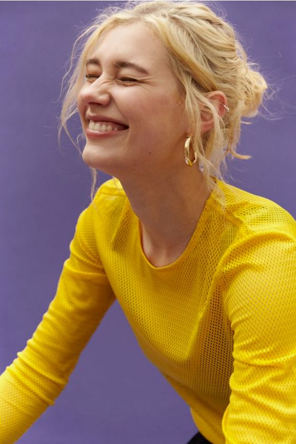 the image is a closeup studio portrait of a young, blonde woman with her hair styled in a loose bun. she is wearing a bright yellow, longsleeved mesh shirt and gold hoop earrings.  she is laughing heartily, showing her teeth, and her expression is one of genuine joy. the background is a solid purple. the overall style is casual and expressive, highlighting the model's happiness.