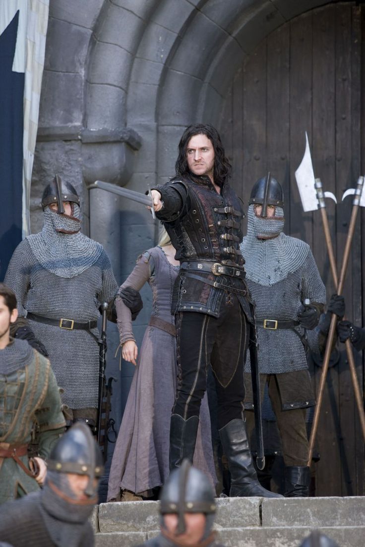 a man with long black hair, clad in dark leather attire and boots, stands prominently on castle steps. he brandishes a sword, pointing it towards the viewer with a serious expression.  he is flanked by two armored knights holding spears, and behind him stands a woman in a flowing gown.  a crowd of people, some in armor, gather at the foot of the steps, their faces obscured or partially hidden. the scene is set against the backdrop of a stone castle, and the overall lighting suggests an overcast day.