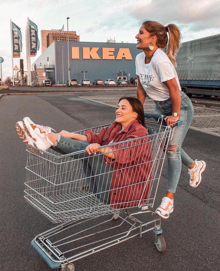 two young women are having fun outside an ikea store. one girl is sitting in a shopping cart while the other girl is pushing it. they are both smiling and looking happy. the girl in the cart is wearing a red jacket and blue jeans, while the girl pushing the cart is wearing a white shirt and blue jeans. they are both wearing white sneakers. the girls are having a great time together and enjoying their friendship.