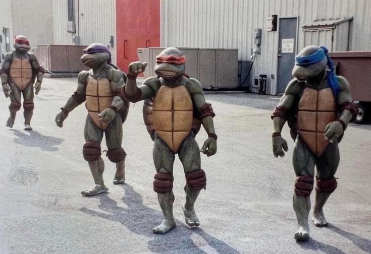 four teenage mutant ninja turtles are walking down a sidewalk.  the turtles are all in their iconic green and brown shell and mask costumes.  they appear to be ready to fight crime.  the one on the far left is leonardo, the leader of the group. he is wearing a blue bandana and is holding two katanas.  the second turtle is michelangelo, who is wearing an orange bandana.  he is holding a pair of nunchakus.  the third turtle is donatello, who is wearing a purple bandana.  he is holding a pair of sai.  the turtle on the far right is raphael, who is wearing a red bandana.  he is holding a pair of katanas.