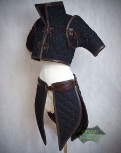 a black quilted gambeson with brown leather accents is displayed on a mannequin. the gambeson features a high collar, short sleeves, and a belted waist with tassets that extend down the thighs. the background is a textured gray wall, and the lighting is soft and even, highlighting the details of the garment.