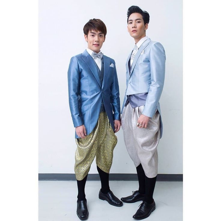 two men in traditional thai attire, standing side by side. the man on the left is wearing a blue suit jacket over a gold patterned pant. the man on the right is wearing a blue suit jacket over a silver pant with a pattern. both men are wearing black shoes.
