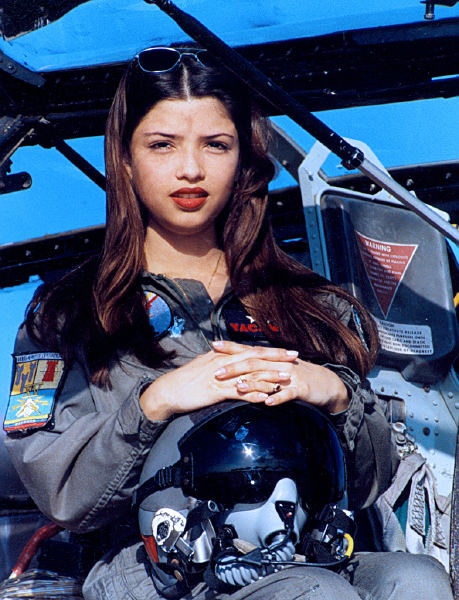 a young female pilot sits in the cockpit of an aircraft. she is wearing a flight suit, sunglasses, and a helmet. she is looking at the camera with a serious expression on her face. she has long, dark hair. she is holding her hands together in her lap. the image is taken from a low angle, looking up at the pilot.