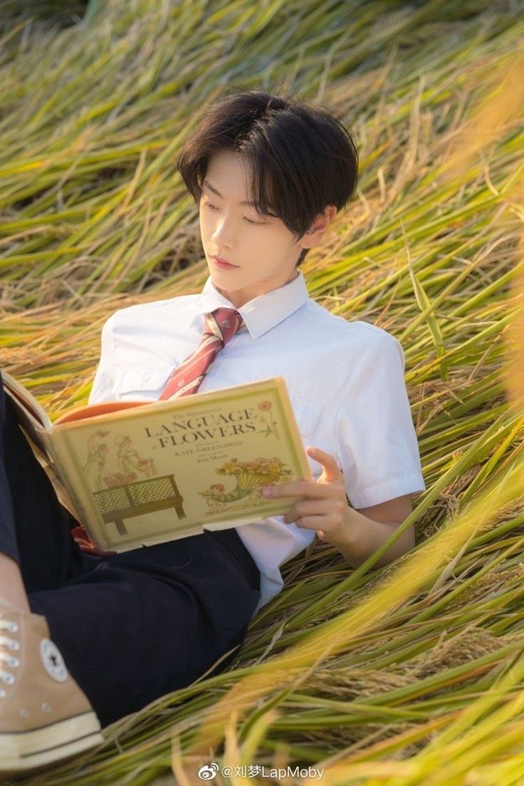 a young asian man is engrossed in a book titled "language of flowers" while relaxing in a golden field, likely a rice paddy. he's dressed in a white shirt and dark pants, with a maroon tie adding a touch of formality. his casual pose and the soft, warm lighting contribute to the serene and peaceful atmosphere of the scene.