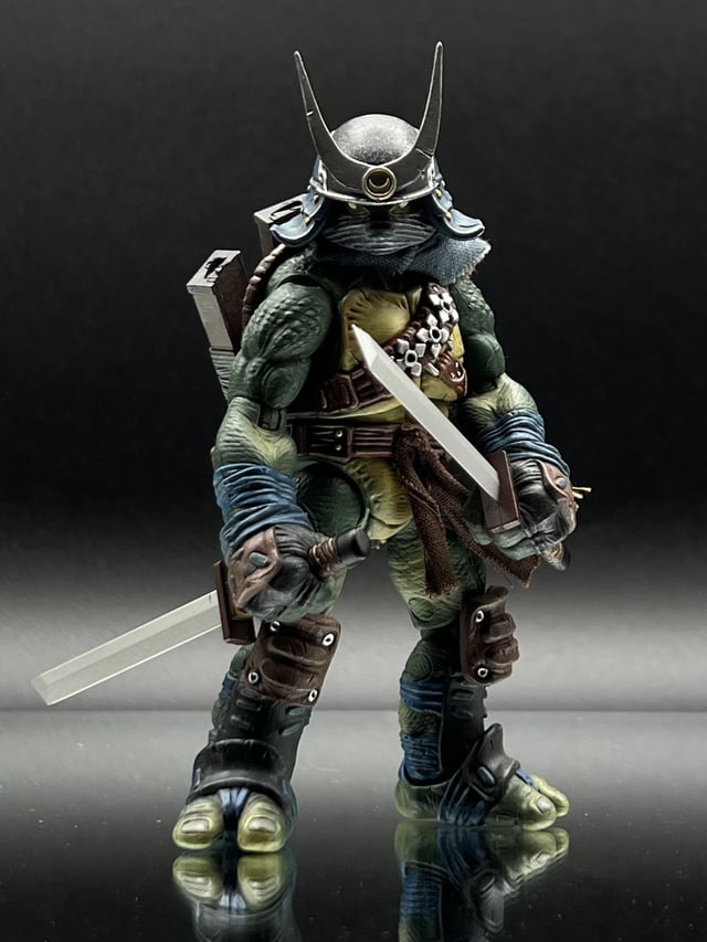 an action figure of a ninja turtle in samurai attire is standing with katana swords in each of its hands.