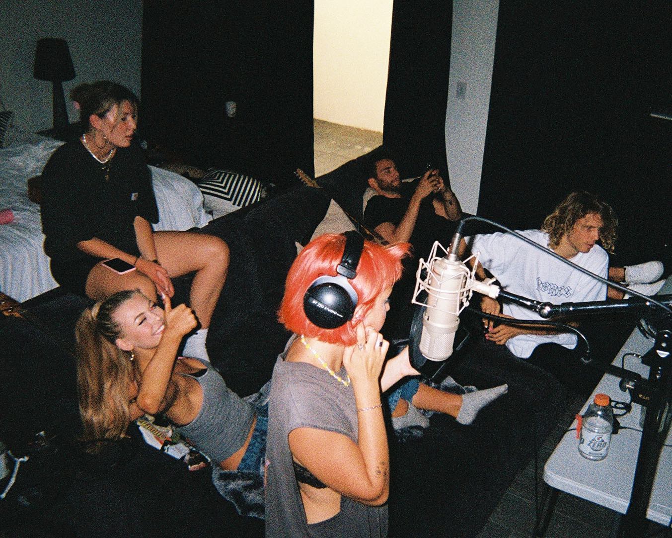 a group of people are gathered in what appears to be a recording studio or creative space. several individuals are sitting on a darkcolored sofa, while one person with bright orange hair and headphones stands close to a microphone, possibly about to record. a woman lying on the floor gives a thumbsup, and another woman sits on the edge of a bed in the background, holding a digital device. a man is using a digital device. the atmosphere is relaxed and informal. the background includes studio equipment, a lamp, and a table with a water bottle.