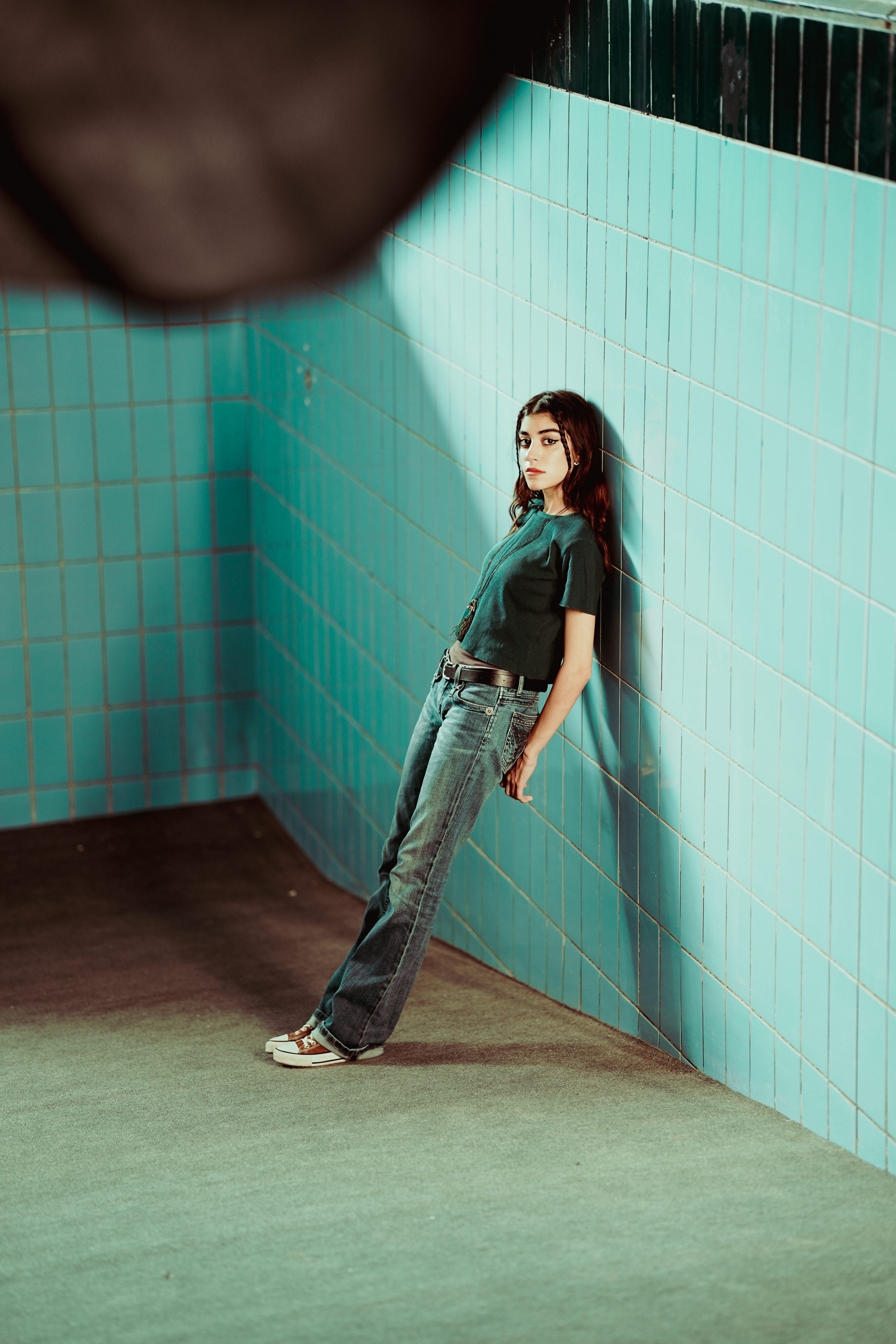 a young woman is leaning against a blue tile wall in a corner of a room. she is wearing jeans, a dark green tshirt, and sneakers. she has brown hair and is looking at the camera.