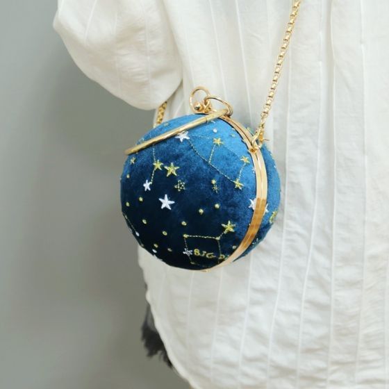 a closeup shows a blue, spherical handbag hanging from a gold chain strap. the bag is decorated with celestialthemed embroidery, featuring stars, constellations, and gold accents. it's presented against the backdrop of a white, vertically striped shirt.