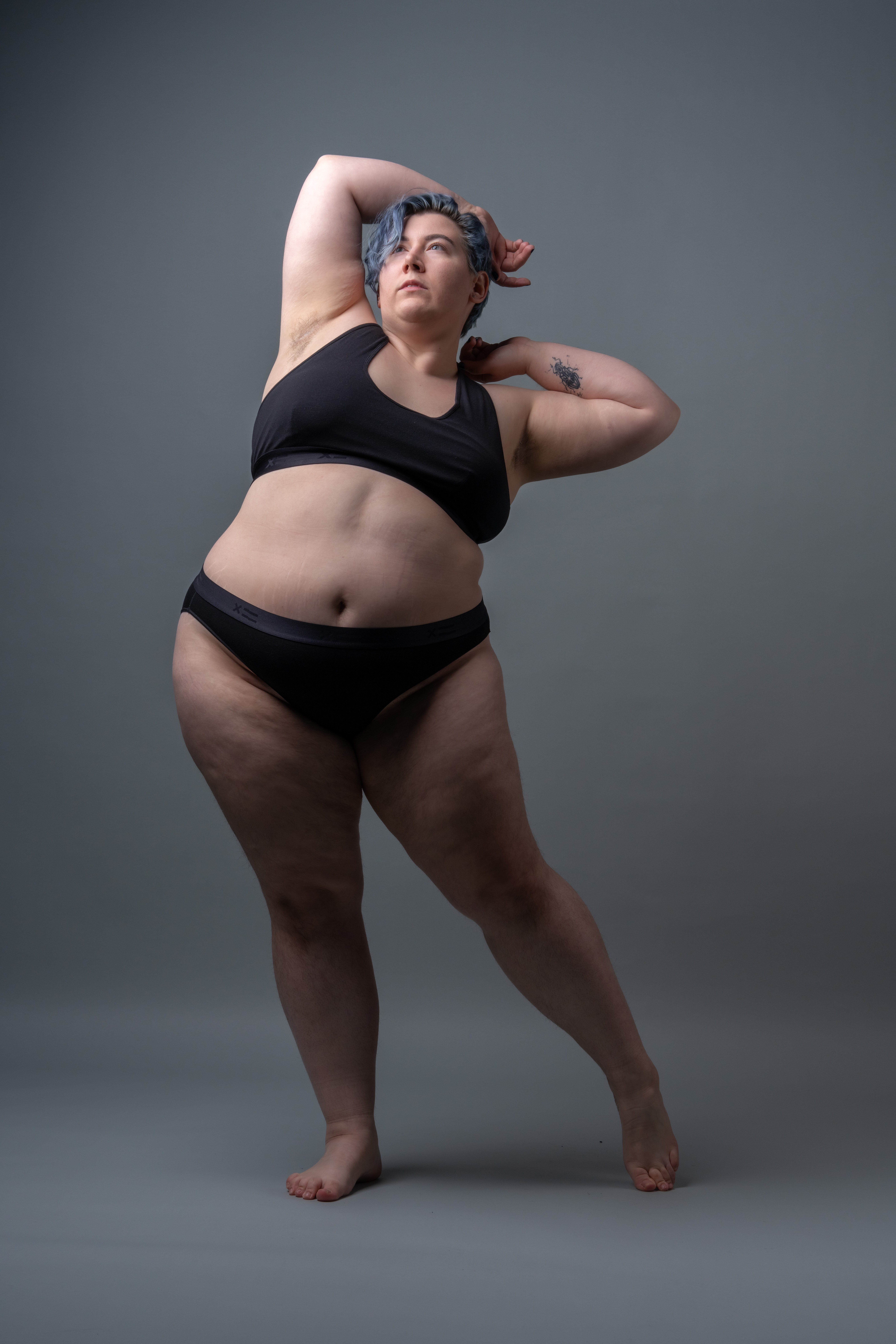 a fulllength studio shot shows a plussize woman with short blue hair posing in a black sports bra and matching underwear against a plain gray background. she has a tattoo on her left arm and is barefoot. her pose involves her arms raised behind her head, with her gaze directed upwards.