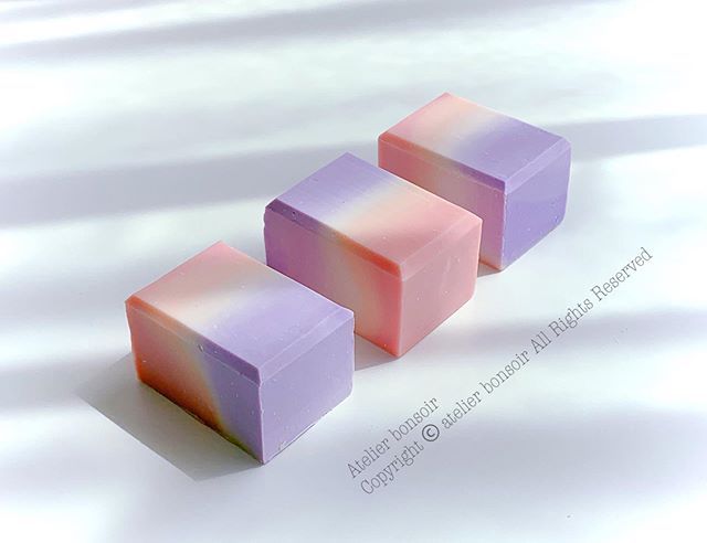 three handmade soap bars are stacked on top of each other. the bottom bar is a soft purple with a light pink on the bottom half, the middle bar is pink with a touch of orange and the top bar is a light purple with an almost white gradient. the soap bars are all handmade, creating a beautiful organic feel.