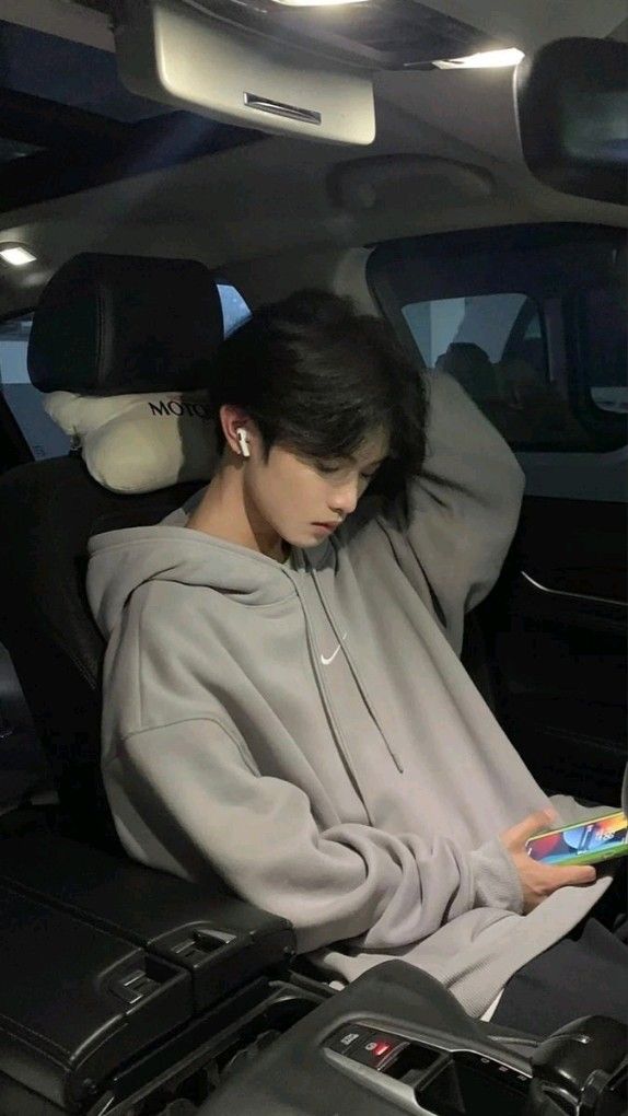 a young asian man with black hair is sitting in the driver's seat of a car. he is wearing a grey hoodie and white headphones, and he is looking down at his smartphone. the car interior is dark and the lighting is soft. the image is taken from an eyelevel perspective.