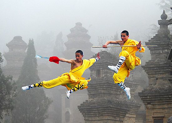 two chinese monks in yellow robes are performing martial arts in front of a foggy mountainous background with ancient temples. one monk is holding a staff and the other is wielding a sword.  the monks are midair, kicking and jumping through the air, showcasing their incredible skills and strength. the image is full of drama and color, and captures the beauty and serenity of the chinese mountains.