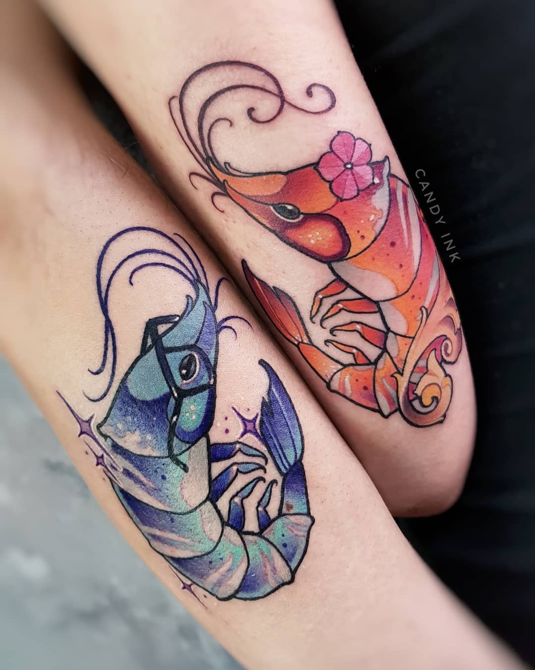 this is an image of two forearms with matching shrimp tattoos on the inner arm. the top arm has a red and orange shrimp with a pink flower on its head, and the bottom arm has a blue and purple shrimp with round glasses on its face. both tattoos are in a neo traditional style and appear to be freshly done.