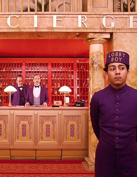 a young man in a purple uniform stands with a stern face behind the counter of a grand hotel's lobby, facing the camera. the counter is made of wood and is ornate. the hotel's sign above the counter says "cier" in gold letters. behind the young man are two men in suits, one is wearing a red tie and the other is wearing a white shirt and a red tie. the room behind the counter has red walls and wooden doors with brass knobs. the door to the right of the counter is open and reveals a hallway. there is a large, goldpainted pillar in the center of the lobby. the floor is covered in a red and gold patterned rug. the entire image has a retro feel.