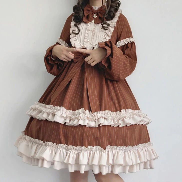 a brown and cream lolita fashion dress with a tiered ruffle skirt and long sleeves. the dress has a high collar and is tied at the waist with a matching brown ribbon. the model is also wearing a brown ribbon in her hair. the dress is very detailed and has a vintage feel.