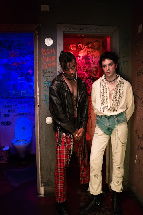 two young men in stylish punk/grunge attire pose in a dimly lit restroom. one wears a black leather jacket and red plaid pants with chains, while the other wears a white ruffled shirt and offwhite jeans, also adorned with chains.  the bathroom walls are covered in graffiti.  the overall aesthetic is edgy and cool, reflecting a rebellious and alternative fashion sense.