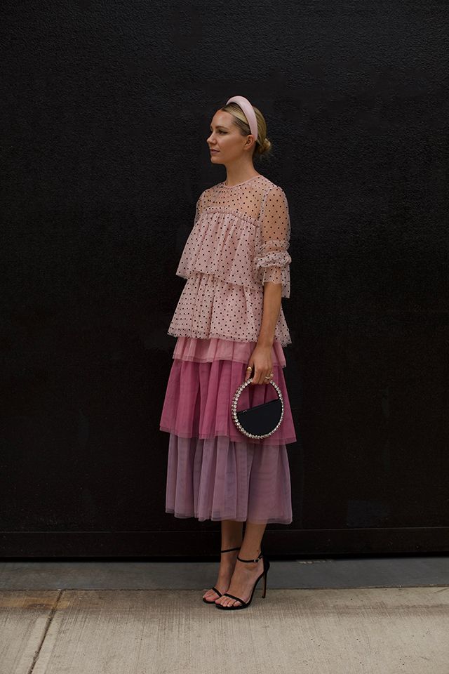 a fulllength shot of a woman wearing a beautiful pink tiered tulle dress with polka dots on the top layer. the dress has a ombre effect, transitioning from light pink to mauve. she's accessorized with a pale pink headband, a small black handbag with a beaded circular frame, and black highheeled sandals. the woman is posing against a black wall, outdoors, possibly in a city setting.  her style is elegant and summery.