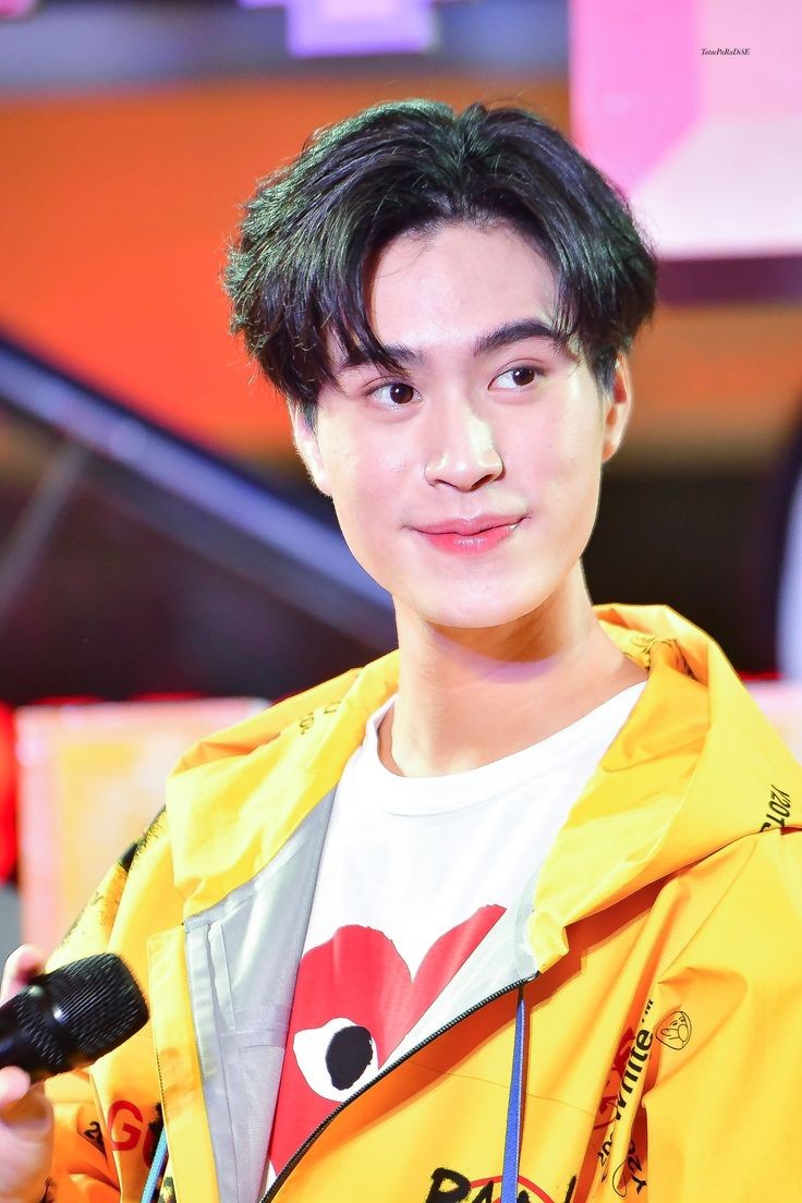a photo of bright vachirawit, a popular thai actor. he is wearing a yellow jacket over a white shirt with a red heart design. he has short, dark hair and is smiling slightly while looking off to the side. he is holding a microphone in his hand, suggesting he's at a public event or fanmeeting. the background is blurry and warmtoned, indicating an indoor setting.