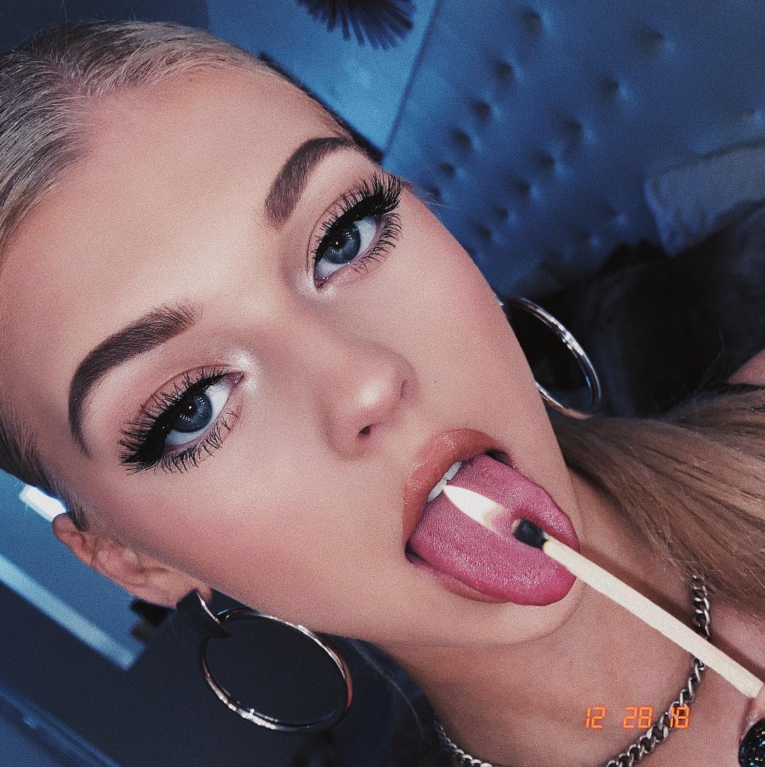 a closeup portrait of a blonde woman with blue eyes and heavy makeup, sticking her tongue out and holding a lit match in her mouth. she is wearing large hoop earrings and a silver chain necklace. a date, "12 28 18," is visible in the bottom right corner of the image.