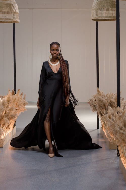 a black woman with long braids is showcasing a stylish long black dress with a high slit. she's wearing a pearl necklace and beige heels, and she's walking down a runway with neutralcolored pampas grass decor on either side.  the setting appears to be an indoor fashion show or photoshoot. the overall aesthetic is elegant and sophisticated.
