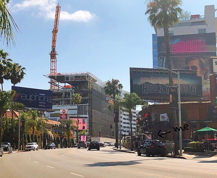 this is a photograph taken in hollywood, california, showing a bustling street scene. a large construction crane is visible in the background, next to a building under construction. prominently displayed are billboards advertising the movie "once upon a time in hollywood" and the hbo show "euphoria."  palm trees line the streets, which have various cars traveling along them. there is also a restaurant visible on the right side of the image, and the person who took the picture has marked their location with an arrow and the word "me." the overall ambiance is a typical sunny hollywood street scene, full of activity and iconic imagery.