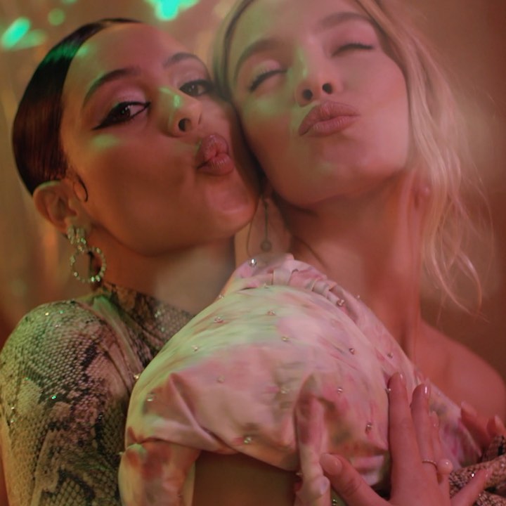 the image is a closeup shot of two young women intimately embracing and puckering their lips as if for a kiss.  one woman has dark hair styled back and is wearing a snakeskinpatterned top; the other has long blonde hair. both women are softly lit with colorful bokeh lighting in the background suggesting a party or nightlife setting.  their expressions are affectionate and sensual. they are holding a piece of light pink clothing, possibly a piece of clothing one of them is wearing. the overall mood is intimate and playful.