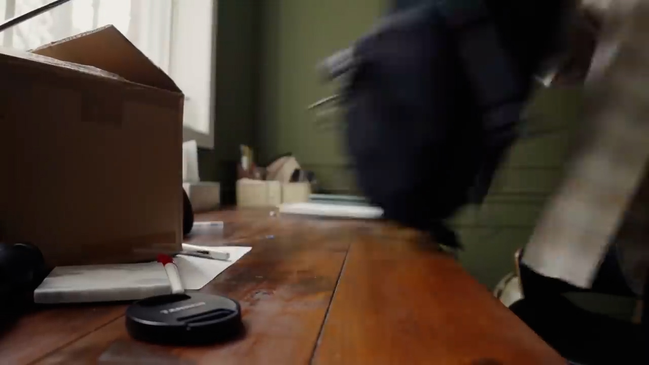 the image shows a cluttered wooden desk with a person holding a dark backpack moving past it. the person is out of focus, suggesting movement, and only their legs and backpack are visible.  the desk is covered with various items including a cardboard box, papers, writing utensils, and a camera lens cap. the lighting is soft and natural, likely coming from a window out of frame. the overall atmosphere suggests a busy and somewhat chaotic moment, perhaps someone leaving or arriving.