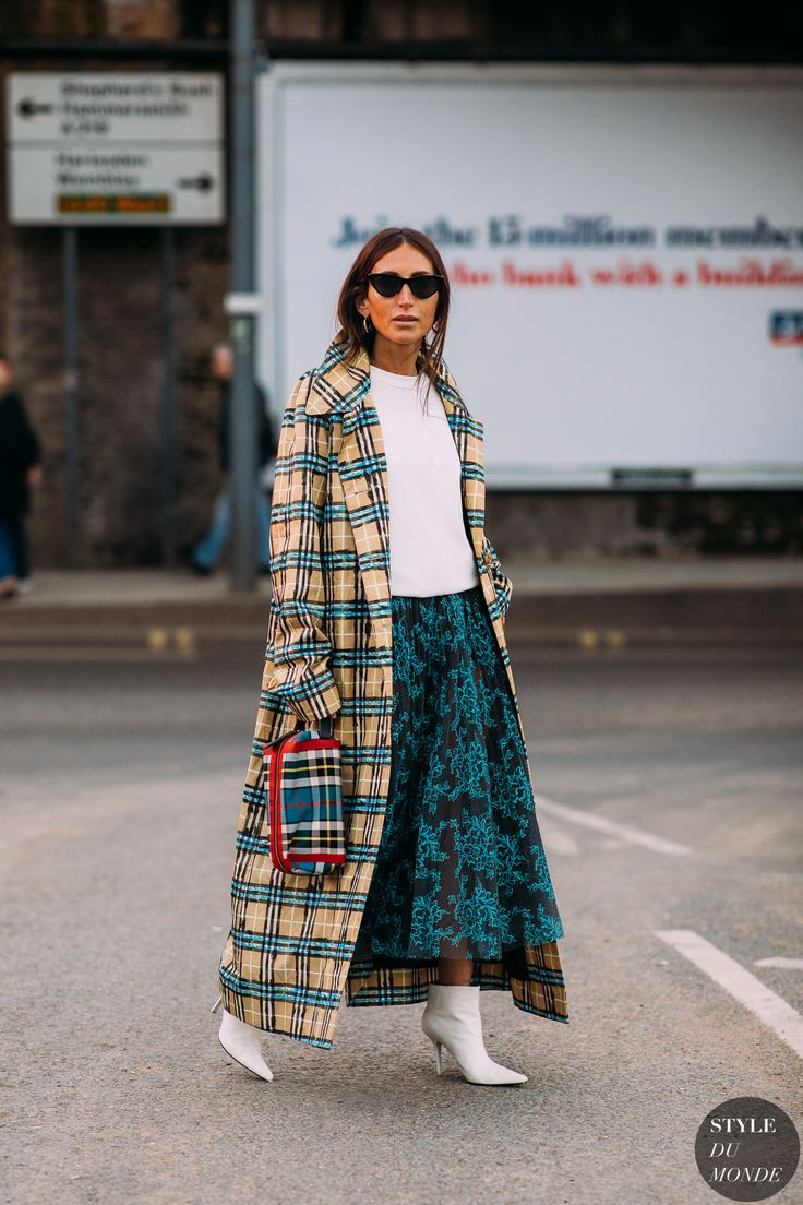 a stylish woman is captured in a fulllength street style shot. she's wearing a beige and brown plaid maxi coat over a white sweater and a dark teal floral midi skirt.  she carries a small plaid crossbody bag and is wearing white ankle boots. her sunglasses add a touch of sophistication to the outfit. the background features a city street with a large billboard, enhancing the urban fashion context. the overall aesthetic is chic, sophisticated and fall/winter appropriate.