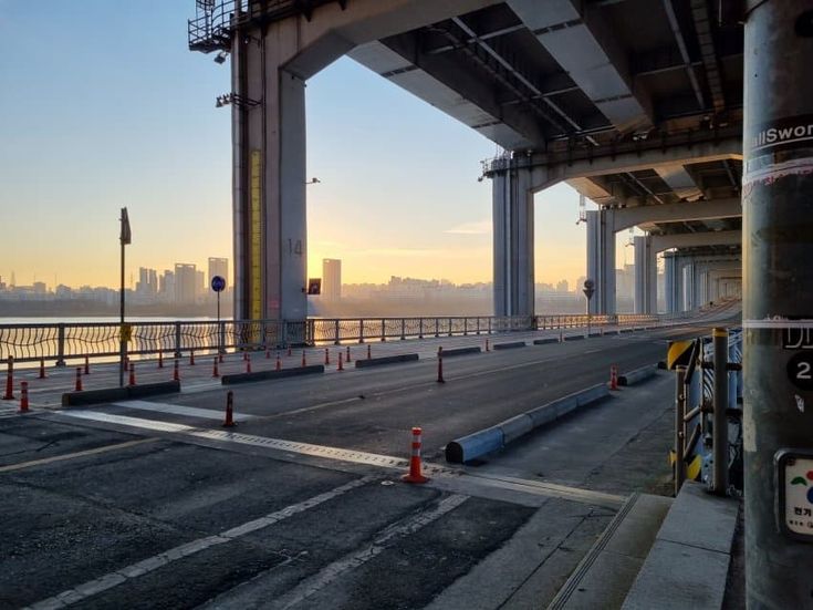 the image shows a wide shot of a bridge in a city at sunrise. the bridge is large and modern, with concrete pillars and a steel structure. the road on the bridge is empty, and the sun is rising over the river in the background. the city skyline is visible in the distance. the overall atmosphere is calm and peaceful. the image is wellcomposed, with the bridge leading the viewer's eye to the sunrise.  there are traffic cones visible on the bridge deck, suggesting some sort of maintenance or construction activity may be taking place.