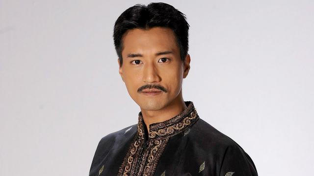 the image is a portrait of a man, likely an actor, against a plain white background. he has short black hair, brown eyes, and a mustache. he is wearing a black shirt with intricate gold embroidery. the lighting is soft, creating a flattering and professional look.