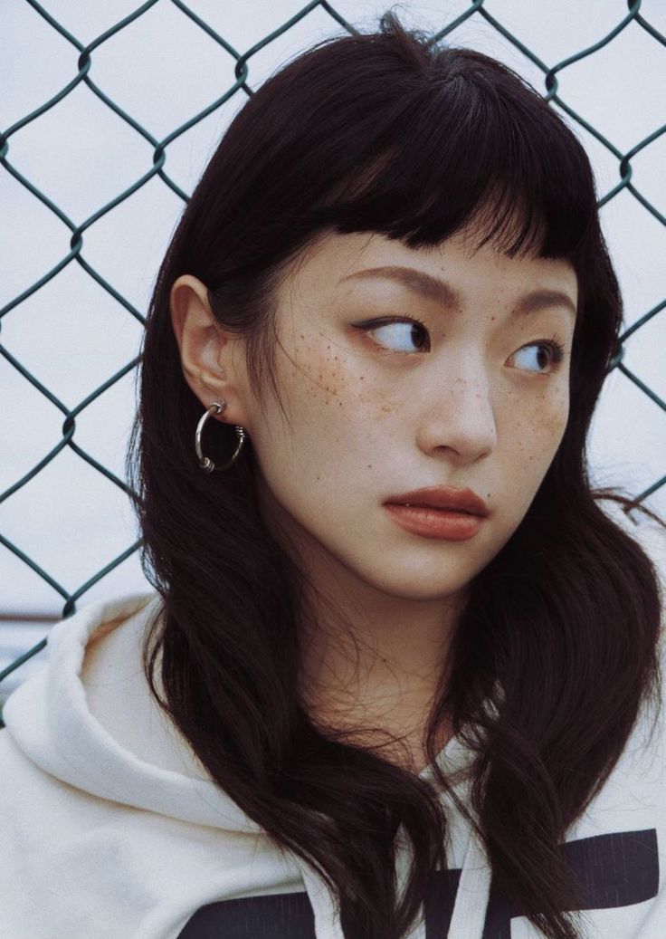 closeup portrait of a young woman with dark hair and bangs, wearing hoop earrings and a white hoodie. she has freckles on her face and is standing near a chain link fence. the image is styled in a casual, natural way with soft lighting.