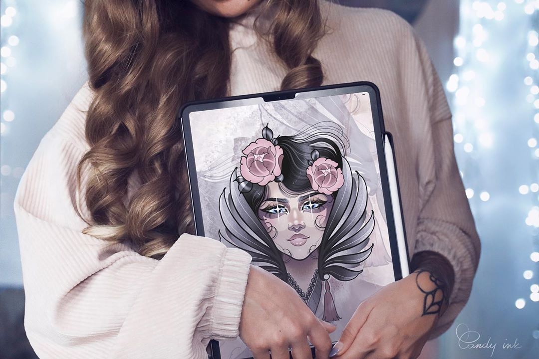 a young woman with long brown hair and a white sweater holds a tablet displaying a digital drawing of a woman's face with angel wings and roses in her hair. the style of the drawing appears to be inspired by neotraditional tattoo art. the background is blurred, suggesting an indoor setting with soft, artificial lighting that creates a cozy atmosphere.