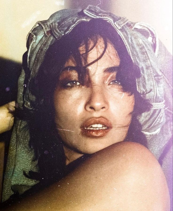 a closeup portrait of a woman with dark hair and denim material draped over her head like a headscarf. she has bangs and a slightly opened mouth. her right shoulder is bare. the image has a vintage filter applied, with visible film grain and color cast.