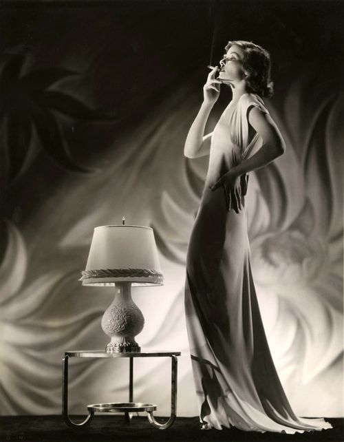 a glamorous woman in a long, flowing dress stands in a studio, smoking a cigarette. she is posing with her hand on her hip, and her head is tilted back as she exhales a plume of smoke. there is a table with a lamp on it in the foreground. the image is in black and white, and the lighting is dramatic, creating strong shadows and highlights. the woman’s silhouette is striking, and her pose is both seductive and elegant.