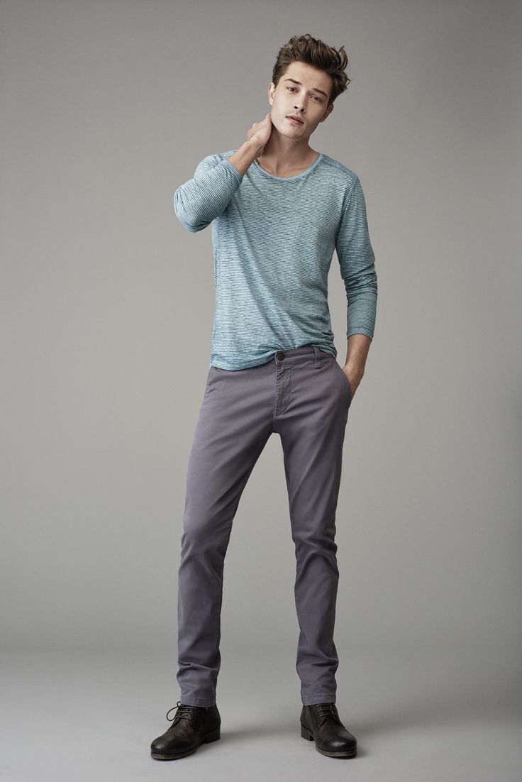 a fullbody shot of a stylish man standing against a neutral grey background. he is wearing a light blue longsleeve shirt, grey trousers, and brown dress shoes. he has one hand raised to his neck and is looking directly at the camera.