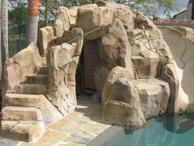 a rocky grotto featuring stone steps leading to a cavelike entrance next to a swimming pool. the structure is composed of artificial rocks and creates a naturalistic water feature in a backyard setting.