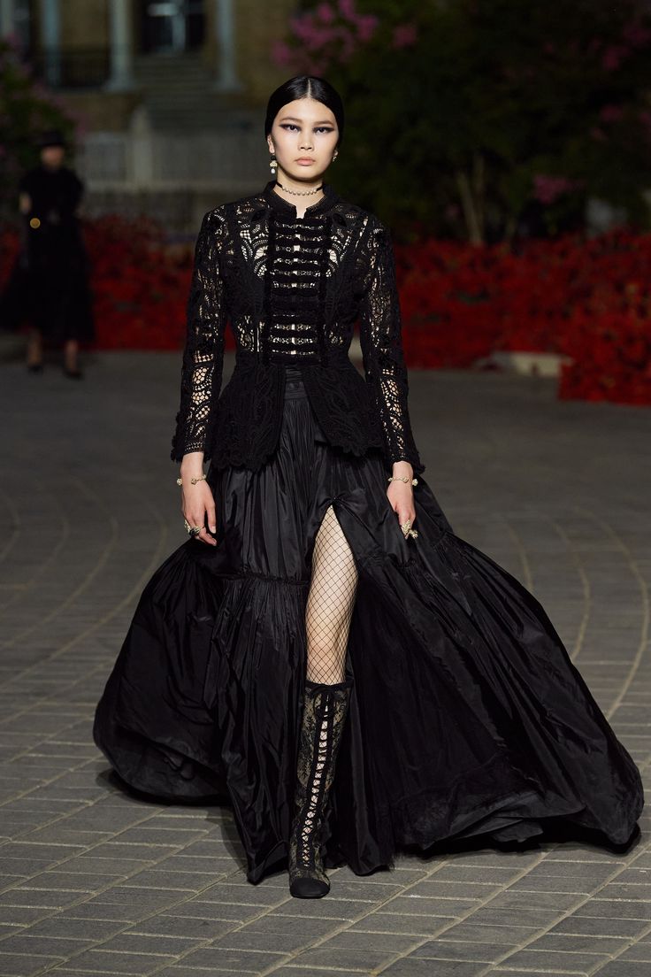 a model walks down a runway wearing a long black dress. the dress is made of lace and features a high slit. she is also wearing black high boots and fishnet stockings. the model has dark hair and makeup. the background is blurry but shows a paved area with red flowers.