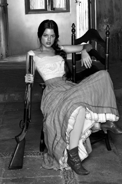 a black and white portrait of a young woman sitting on an old wooden chair, holding a shotgun in her right hand, with a serious expression, wearing a long dress and boots.