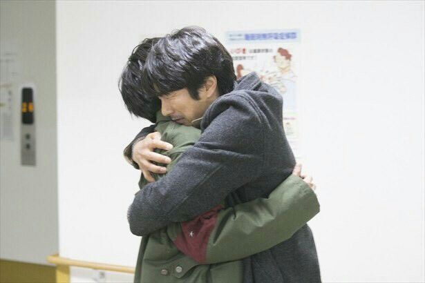 two men are hugging each other tightly. the man in the back is wearing a grey jacket and the man in the front is wearing a green jacket. the man in the back is looking down, and the man in the front is looking up. they are both looking sad and emotional.  there is a white wall in the background.