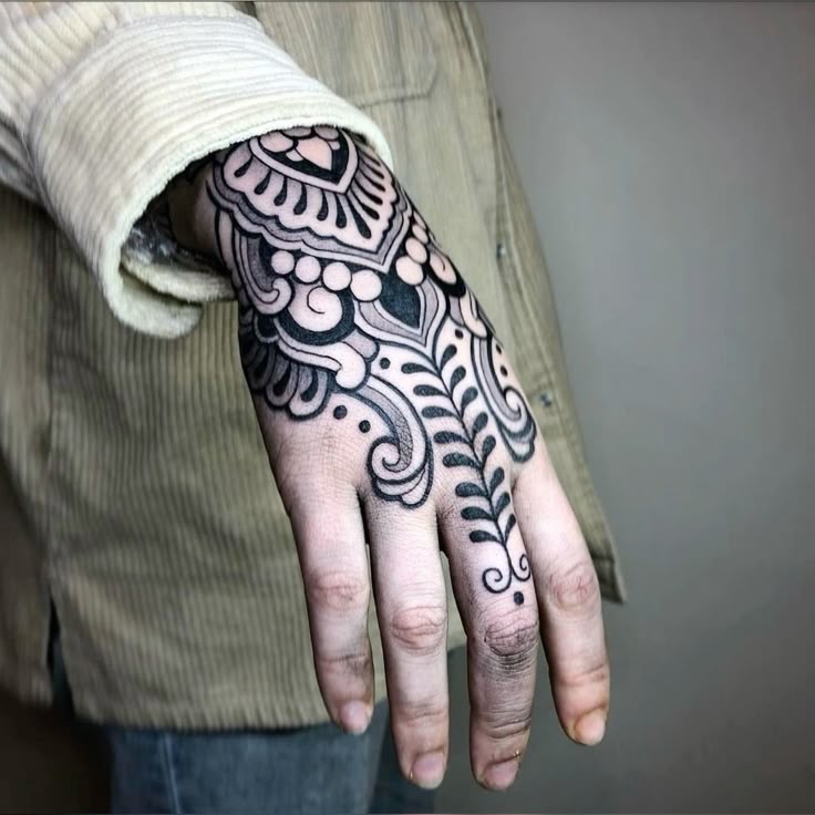 a closeup shot of a person's hand with a detailed black and white tattoo. the tattoo covers the back of the hand and extends onto the fingers. the design features intricate patterns, including floral and geometric elements. the person is wearing a longsleeved corduroy shirt, with the sleeve partially rolled up to reveal the tattoo. the background is a plain wall, drawing focus to the hand and tattoo.
