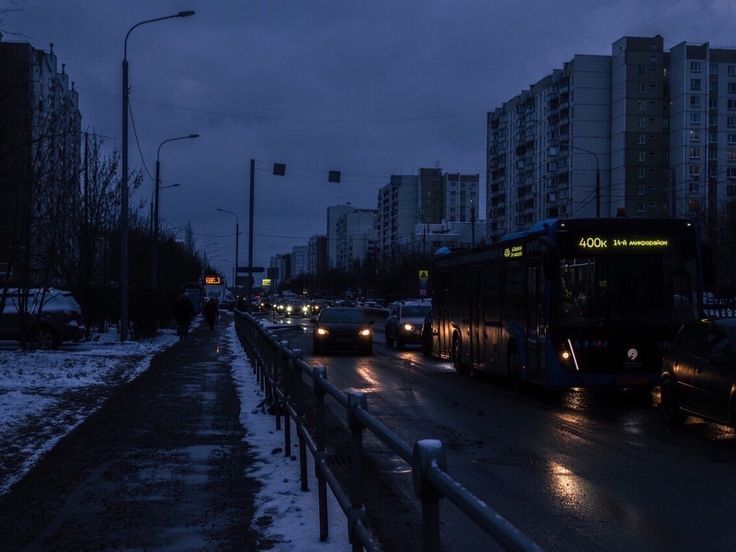 the image depicts a city street at night during winter.  it's dark and cold, with snow on the ground and sidewalks. a bus is the central focus, moving along the street with other cars.  residential buildings line the street, creating a moody and gloomy atmosphere. the scene is dimly lit by streetlights. the overall feeling is one of quiet urban solitude.