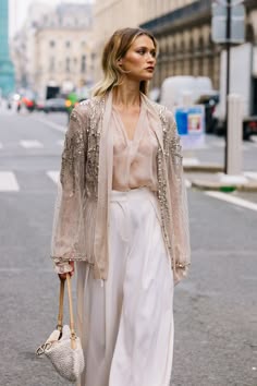 a young woman in a white outfit and a sequin jacket walks down a city street. she has blonde hair and is wearing a white top and white pants. she is carrying a white handbag.