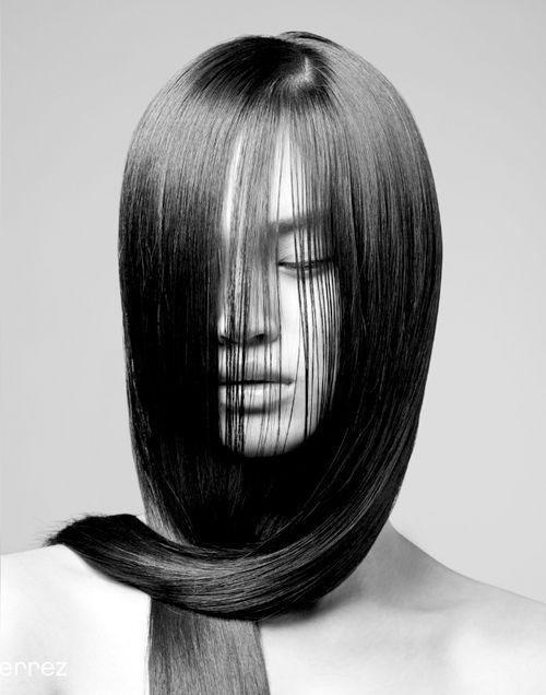 a woman with long black hair, styled in a sleek, straight style, covering most of her face. the image is in black and white, creating a dramatic and mysterious mood. she is looking down, creating a sense of introspective thought or contemplation. the light highlights her sharp cheekbones and the perfect symmetry of her face. the simplicity of the background and the dramatic style of the hair make this image very striking and alluring.